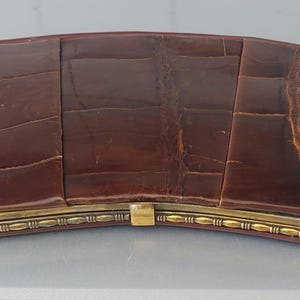 Könnte beinhalten: Eine geschwungene, rechteckige braune Clutch mit glänzendem, texturiertem Krokodilleder-Muster. Das Accessoire verfügt über einen goldfarbenen Metallrahmen mit einem dekorativen, geprägten Muster am Rand und einen mittigen Verschluss.