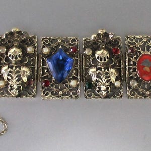 May include: A vintage gold-toned bracelet with six rectangular panels. Each panel features intricate filigree and is adorned with various gemstones, including a blue, red, and green stone, and a cameo. A delicate chain secures the bracelet.