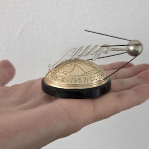 Puede incluir: Un modelo metálico del satélite Sputnik con una esfera plateada y cuatro antenas finas, montado sobre una base semiesférica dorada con un mapa grabado y el texto 4-X-1957r CCCP, todo sobre una base circular de color negro.