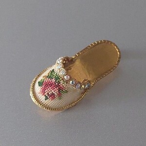 May include: A white slipper with a gold trim and a pink embroidered flower design. The slipper is decorated with rhinestones.