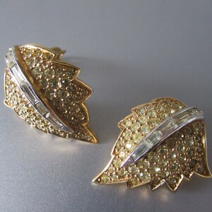 May include: A pair of gold tone leaf-shaped earrings with clear rhinestone accents. The earrings feature a row of baguette-shaped rhinestones in the center of each leaf.