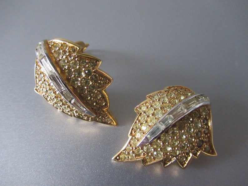 May include: A pair of gold tone leaf-shaped earrings with clear rhinestone accents. The earrings feature a row of baguette-shaped rhinestones in the center of each leaf.