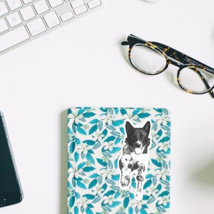 May include: Black and white illustration of a dog running, printed on a white sticker. The sticker is placed on a blue and white floral notebook.
