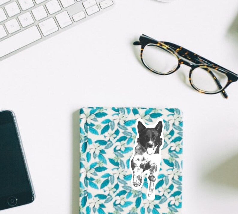 May include: Black and white illustration of a dog running, printed on a white sticker. The sticker is placed on a blue and white floral notebook.
