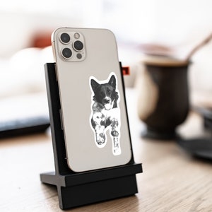 May include: A black and white sticker of a dog with a white chest and paws, on a black phone case. The phone case is attached to a black stand.