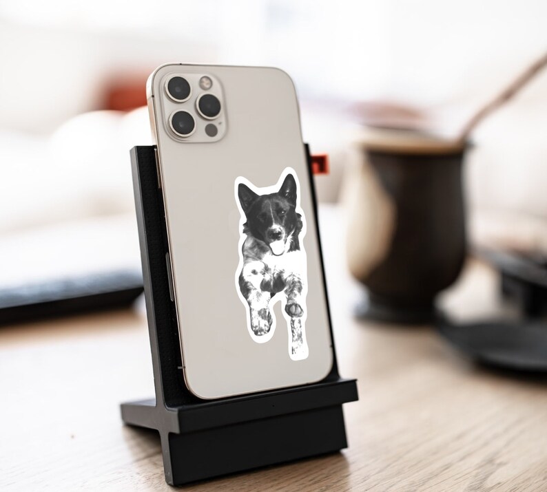 May include: A black and white sticker of a dog with a white chest and paws, on a black phone case. The phone case is attached to a black stand.