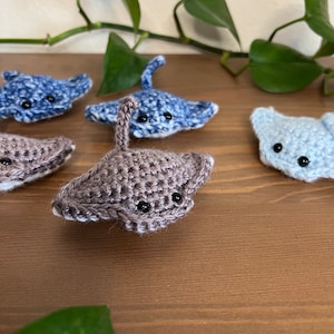May include: Four crocheted stingray plushies in shades of blue and gray. The plushies have black eyes and are arranged on a wooden surface.