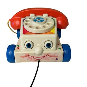 May include: A vintage Fisher-Price toy phone with a red handset, a white face with blue eyes, and a rainbow dial. The phone has blue wheels and a pull string.
