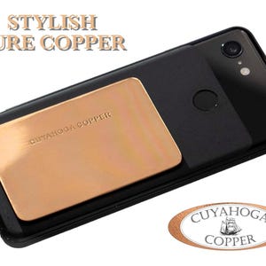 May include: A black smartphone with a copper backplate that says "CUYAHOGA COPPER". The phone has a dual camera lens and a fingerprint sensor. The phone is on a white background with a copper oval sticker that says "CUYAHOGA COPPER" with a ship graphic.