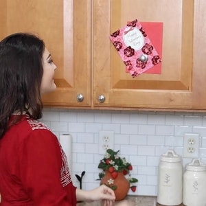 May include: A Valentine's Day card with a floral design is attached to a wooden cabinet. The card has a pink background with red roses and the words "Happy Valentine's Day". A woman in a red shirt is in the foreground.