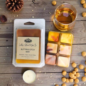 Peut inclure: Un emballage en plastique transparent contenant un fondant parfumé au beurre et à la bière, avec une étiquette indiquant "Lotus Street Wax Co. BUTTERSCOTCH BEER". Un fondant à six cubes et un verre de liquide ambré sont posés sur une surface en bois.