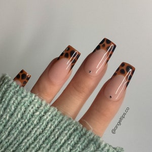 May include: A hand with long, square-shaped artificial nails. The nails feature a French tip design with a brown leopard print pattern. Some nails have small, silver rhinestones. The nails are set against a light green sweater.