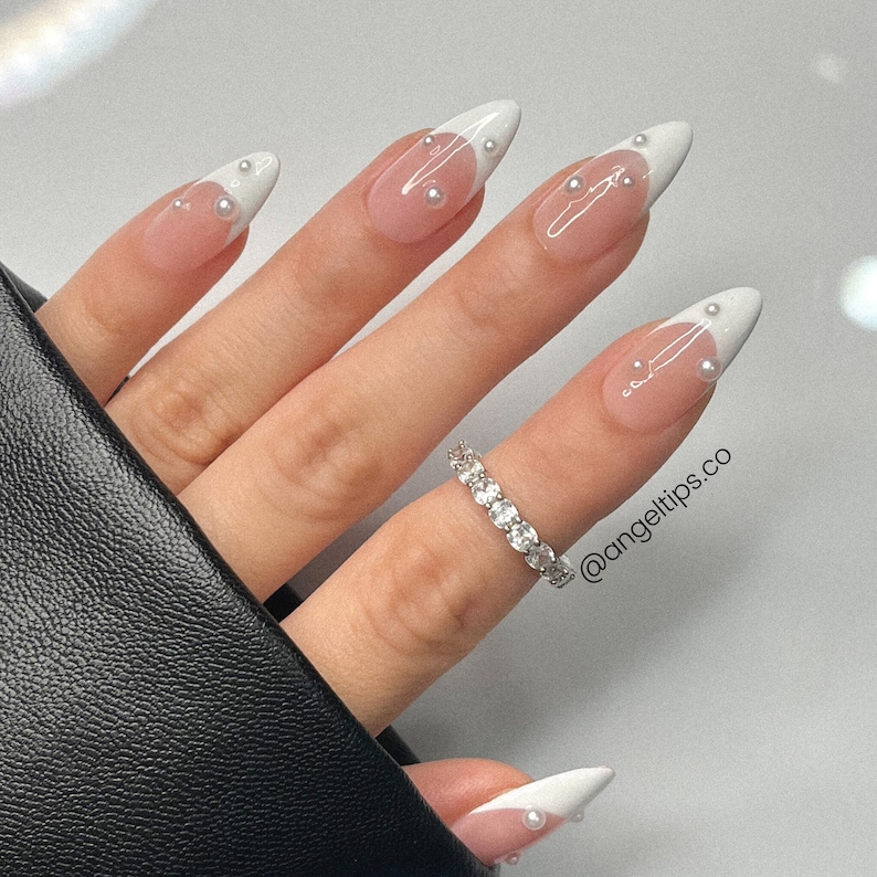 May include: Close-up of a hand with white and nude stiletto nails with pearl accents. The nails have a white tip with a nude base. The hand is wearing a silver ring with small round stones.