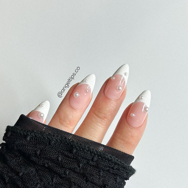 May include: White almond-shaped press-on nails with a white tip and pearl accents. The nails are shown on a hand wearing a black mesh glove. The Instagram handle @angeltips.co is visible in the image.