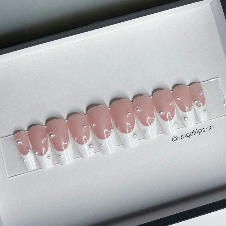 May include: A set of ten pink and white press-on nails with a French manicure design and pearl accents. The nails are arranged in a row on a white surface. @angeltips.co
