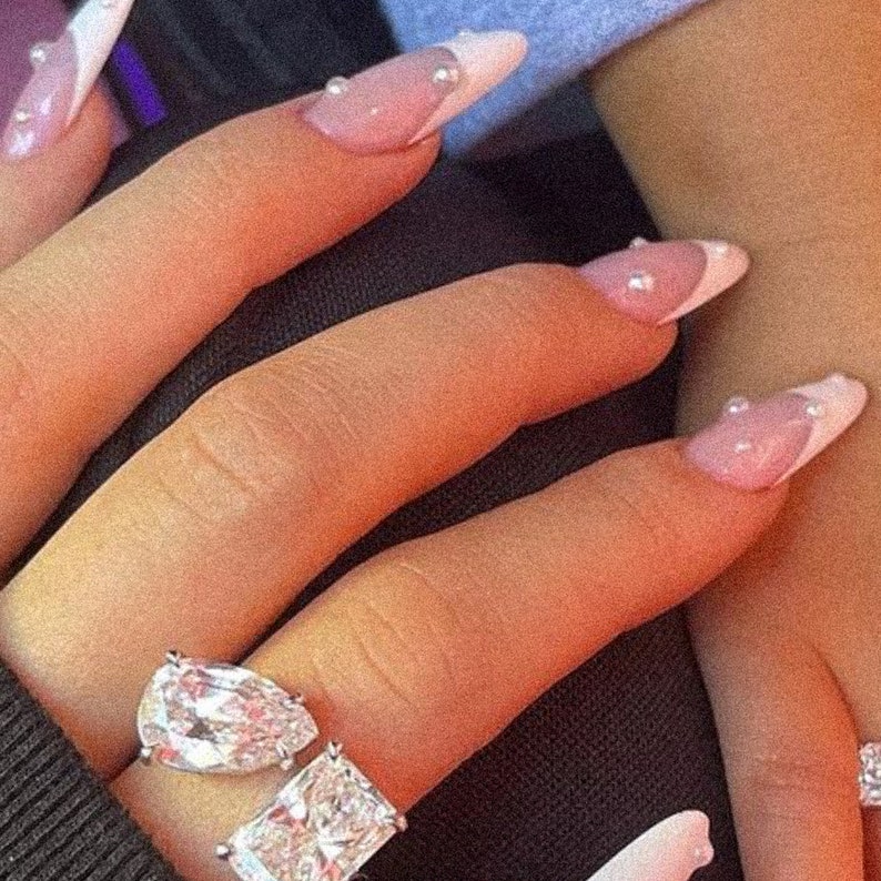May include: A close-up of a hand wearing a large, pear-shaped diamond ring with a smaller, square-shaped diamond ring on the same finger. The hand also has long, white-tipped nails with small pearl accents.
