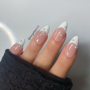 May include: Close-up of a hand with white french manicured nails adorned with small pearl accents. The nails are long and pointed, with a white tip and a nude base. The pearls are scattered across the nails, creating a delicate and elegant look. The hand is wearing a black sweater.