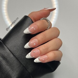 May include: Close-up of a hand with long, almond-shaped nails. The nails are painted with a clear base and a white tip, creating a French manicure. Each nail is adorned with small, white pearl-like embellishments.