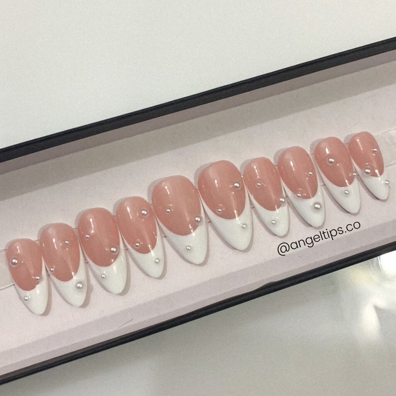 May include: A set of ten false nails with a white french tip design and pearl accents. The nails are in a box with the text "@angeltips.co" on the bottom.