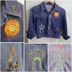 May include: A denim jacket with embroidered patches featuring a smiling sun, a teepee, a headdress, and a cactus with flowers. The jacket is a light blue colour and has a classic style.