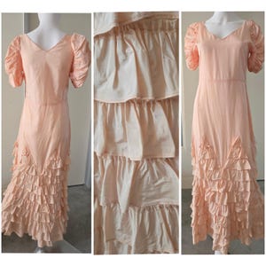 May include: A peach-coloured vintage dress with a V-neckline and short, puffed sleeves. The dress features a tiered, ruffled skirt and a fitted bodice. The fabric appears to be a lightweight material. The dress is displayed on a mannequin.