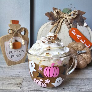 May include: A glass mug with a pink pumpkin and ghost design, filled with a pumpkin spice latte topped with whipped cream and cinnamon. The mug is sitting on a wooden table with fall decorations, including a wooden pumpkin sign that says "Pumpkin Punch" and a white wooden pumpkin with a "Fall" tag.