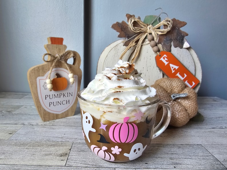 May include: A glass mug with a pink pumpkin and ghost design, filled with a pumpkin spice latte topped with whipped cream and cinnamon. The mug is sitting on a wooden table with fall decorations, including a wooden pumpkin sign that says "Pumpkin Punch" and a white wooden pumpkin with a "Fall" tag.