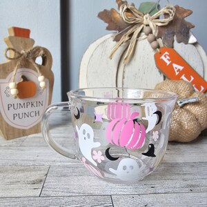 May include: A clear glass mug with a Halloween theme. The mug has pink and white ghosts, black bats, and pink pumpkins. The text "Pumpkin Punch" is visible on a wooden sign in the background.