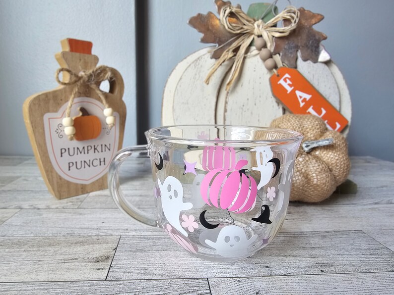 May include: A clear glass mug with a Halloween theme. The mug has pink and white ghosts, black bats, and pink pumpkins. The text "Pumpkin Punch" is visible on a wooden sign in the background.
