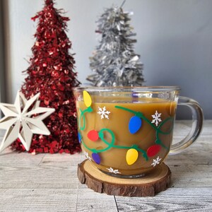 May include: A clear glass mug with a brown liquid inside. The mug has a green vine design with colorful lights and snowflakes. The mug is sitting on a wooden coaster.