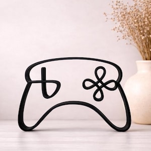 Könnte beinhalten: Schwarze, minimalistische Umrisse eines Videospiel-Controllers. Der Controller hat ein Steuerkreuz und eine Vier-Tasten-Anordnung. Das Design befindet sich auf einer weißen Oberfläche, mit einer Vase mit Trockenblumen im Hintergrund.