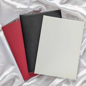 May include: Three rectangular cards in red, black, and white are arranged on a white satin surface. The cards are leaning against each other, with the white card in the foreground. The satin has soft folds.