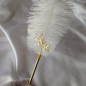May include: A white ostrich feather pen featuring a gold-toned metal shaft and a gold-coloured acrylic monogram attachment displaying the letters Z&R. The quill is accented with a small gold-toned metal collar where the feather meets the pen shaft.