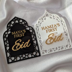 Peut inclure: Deux plaques d&eacute;coratives pour l'A&iuml;d. L'une est noire avec le texte dor&eacute; "Maya's First Eid". L'autre est blanche avec le texte dor&eacute; "Hamza's First Eid". Les deux plaques ont une forme arqu&eacute;e avec des motifs d&eacute;coup&eacute;s.