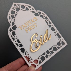 Peut inclure: D&eacute;coration blanche et dor&eacute;e pour l'A&iuml;d Moubarak avec le texte "Yahya's First Eid". La d&eacute;coration a une bordure d&eacute;corative d&eacute;coup&eacute;e et une police dor&eacute;e. Le fond est de couleur grise unie.