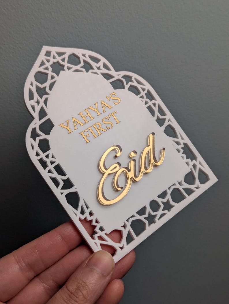 Peut inclure: D&eacute;coration blanche et dor&eacute;e pour l'A&iuml;d Moubarak avec le texte "Yahya's First Eid". La d&eacute;coration a une bordure d&eacute;corative d&eacute;coup&eacute;e et une police dor&eacute;e. Le fond est de couleur grise unie.