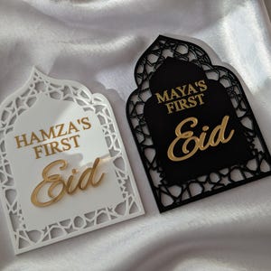 Peut inclure: Deux plaques d&eacute;coratives pour l'A&iuml;d. L'une est blanche avec l'inscription dor&eacute;e "Hamza's First Eid". L'autre est noire avec l'inscription dor&eacute;e "Maya's First Eid". Les deux plaques ont un design en forme d'arche.