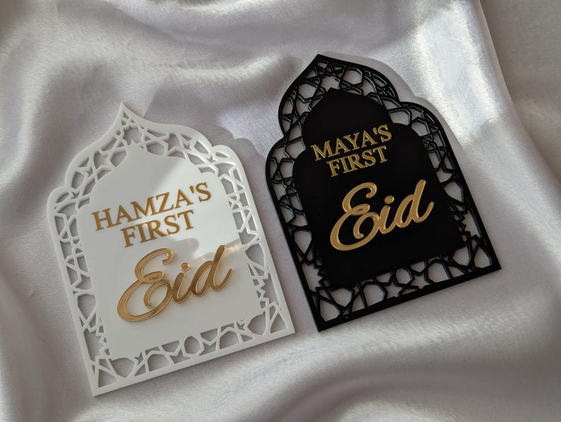 Peut inclure: Deux plaques d&eacute;coratives pour l'A&iuml;d. L'une est blanche avec l'inscription dor&eacute;e "Hamza's First Eid". L'autre est noire avec l'inscription dor&eacute;e "Maya's First Eid". Les deux plaques ont un design en forme d'arche.