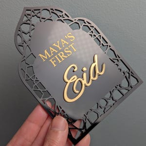 Peut inclure: Plaque d&eacute;corative noire avec un motif g&eacute;om&eacute;trique d&eacute;coup&eacute;. La plaque porte les mots "MAYA'S FIRST Eid" en lettres dor&eacute;es. La plaque est tenue dans une main sur un fond gris.