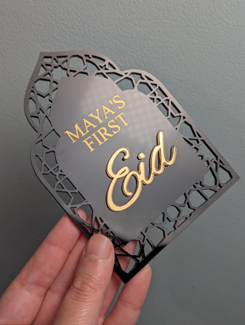 Peut inclure: Plaque d&eacute;corative noire avec un motif g&eacute;om&eacute;trique d&eacute;coup&eacute;. La plaque porte les mots "MAYA'S FIRST Eid" en lettres dor&eacute;es. La plaque est tenue dans une main sur un fond gris.