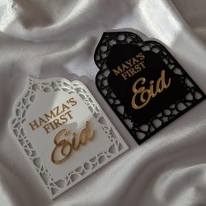 Peut inclure: Deux plaques d&eacute;coratives de l'A&iuml;d, une blanche et une noire, avec des lettres dor&eacute;es. La plaque blanche indique "Hamza's First Eid", et la plaque noire indique "Maya's First Eid". Les deux plaques ont un design d&eacute;coup&eacute; orn&eacute; sur les bords.