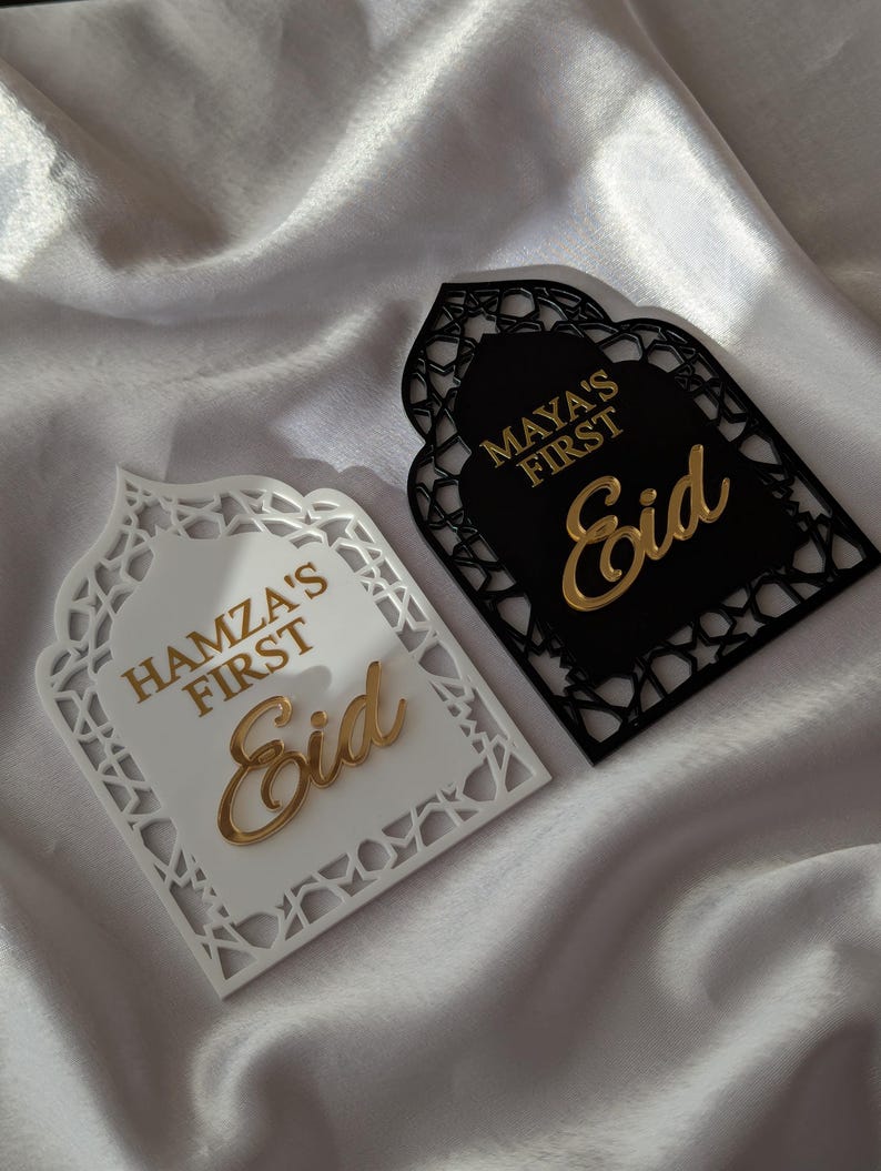 Peut inclure: Deux plaques d&eacute;coratives de l'A&iuml;d, une blanche et une noire, avec des lettres dor&eacute;es. La plaque blanche indique "Hamza's First Eid", et la plaque noire indique "Maya's First Eid". Les deux plaques ont un design d&eacute;coup&eacute; orn&eacute; sur les bords.