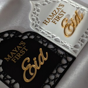 Peut inclure: Deux plaques d&eacute;coratives pour l'A&iuml;d, une blanche et une noire, avec des motifs d&eacute;coup&eacute;s complexes. La plaque blanche porte l'inscription "Hamza's First Eid" en lettres dor&eacute;es, avec un script "Eid" dor&eacute;. La plaque noire indique "Maya's First Eid" avec un script "Eid" dor&eacute;.