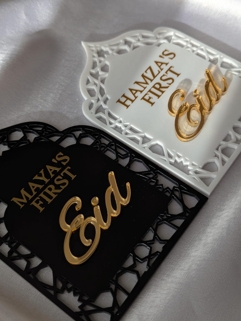 Peut inclure: Deux plaques d&eacute;coratives pour l'A&iuml;d, une blanche et une noire, avec des motifs d&eacute;coup&eacute;s complexes. La plaque blanche porte l'inscription "Hamza's First Eid" en lettres dor&eacute;es, avec un script "Eid" dor&eacute;. La plaque noire indique "Maya's First Eid" avec un script "Eid" dor&eacute;.