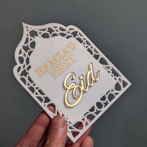 Peut inclure: D&eacute;coration de l'A&iuml;d blanche et dor&eacute;e avec le texte "Hamza's First Eid". La d&eacute;coration a une bordure d&eacute;corative avec un motif d&eacute;coup&eacute;. Le texte est en or et le fond est blanc.