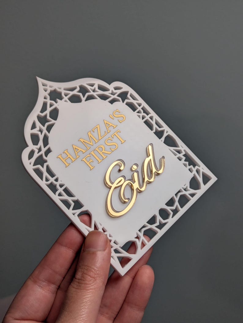 Peut inclure: D&eacute;coration de l'A&iuml;d blanche et dor&eacute;e avec le texte "Hamza's First Eid". La d&eacute;coration a une bordure d&eacute;corative avec un motif d&eacute;coup&eacute;. Le texte est en or et le fond est blanc.