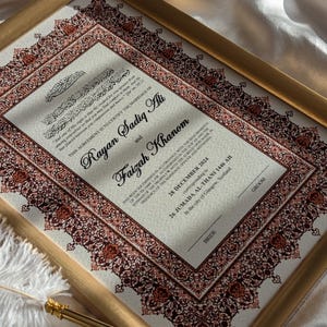 May include: A framed Islamic marriage certificate with intricate red and brown floral designs. The text reads "This agreement is to certify the marriage of Rayan Sadiq Ali and Faizah Khanom." The date of the marriage is 28 December 2024, corresponding to 26 Jumada Al-Thani 1446 AH in the city of Glasgow, Scotland.