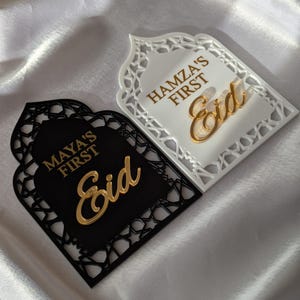 Peut inclure: Deux plaques d&eacute;coratives de l'A&iuml;d, une noire et une blanche, avec des motifs d&eacute;coup&eacute;s complexes. La plaque noire porte l'inscription "MAYA'S FIRST Eid" en lettres dor&eacute;es, tandis que la plaque blanche indique "HAMZA'S FIRST Eid". Sur fond blanc soyeux.