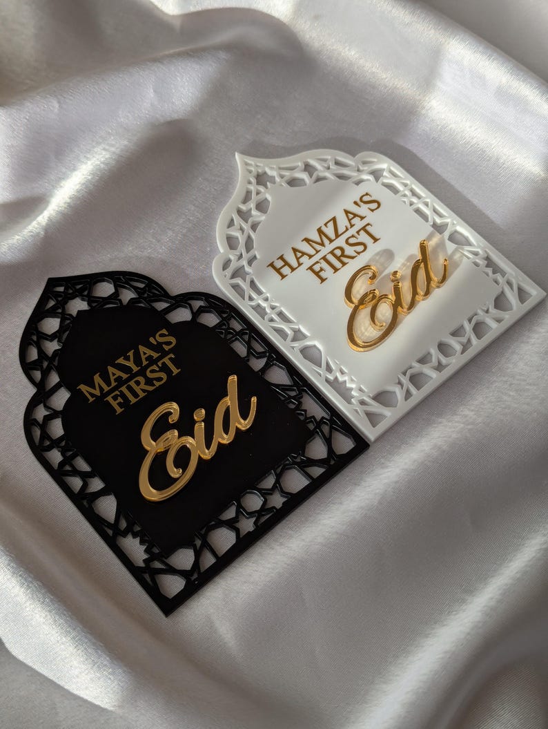 Peut inclure: Deux plaques d&eacute;coratives de l'A&iuml;d, une noire et une blanche, avec des motifs d&eacute;coup&eacute;s complexes. La plaque noire porte l'inscription "MAYA'S FIRST Eid" en lettres dor&eacute;es, tandis que la plaque blanche indique "HAMZA'S FIRST Eid". Sur fond blanc soyeux.
