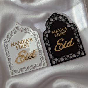 Peut inclure: Deux plaques d&eacute;coratives pour l'A&iuml;d, une blanche et une noire, avec des lettres dor&eacute;es. La plaque blanche indique "Hamza's First Eid", et la plaque noire indique "Maya's First Eid". Les deux plaques ont un design en forme d'arche orn&eacute;.
